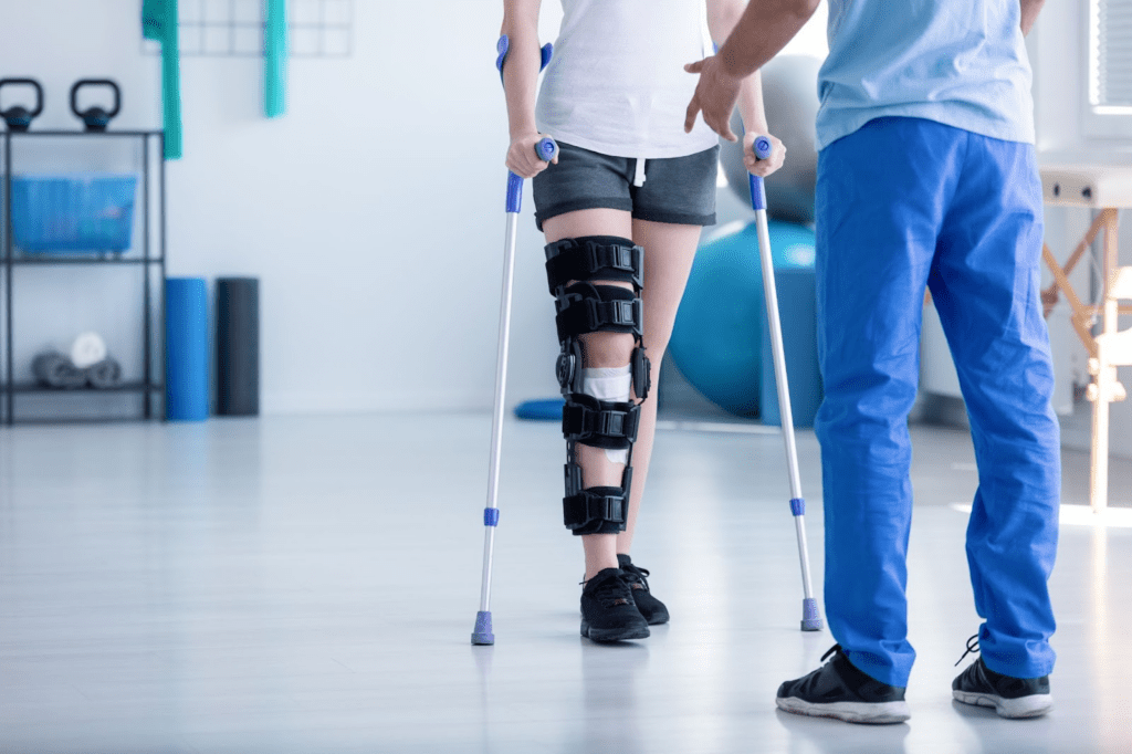 physical therapist in scrubs standing in front of a woman who has had knee surgery walking on crutches during a physical thearpy session