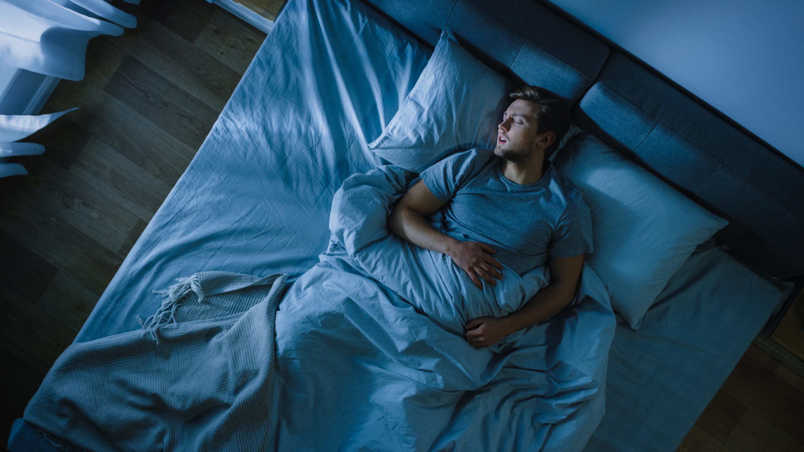 Top View of Young Man Sleeping Cozily on a Bed in His Bedroom at Night showing his posture while sleeping. Blue Nightly Colors with Cold Weak Lamppost Light Shining Through the Window.